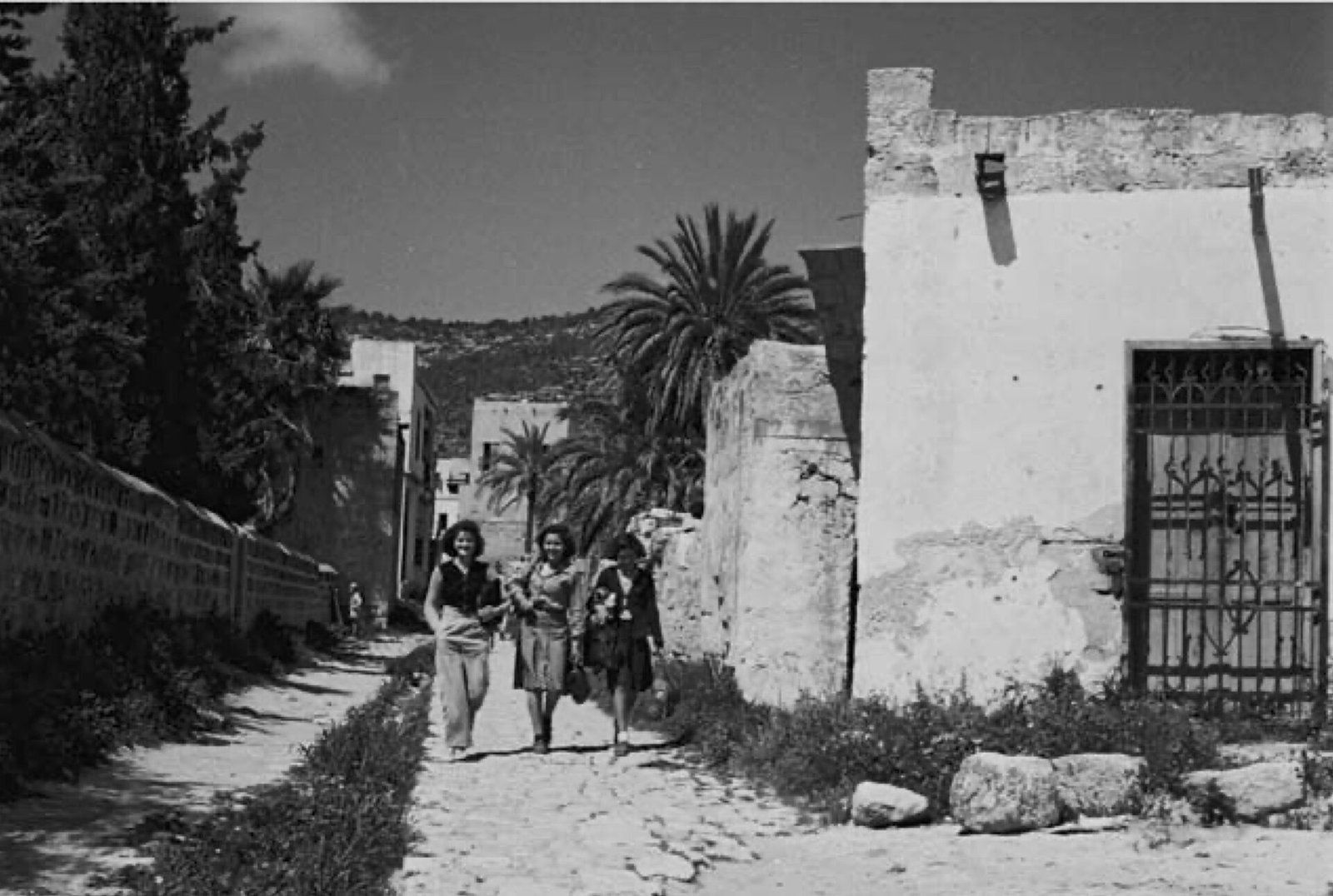 Black and white photo of three young people walking down a stone lane past old village houses in al-Bassa in 1950
