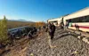 A derailed high-speed train on its side on an embankment with people standing on the track beside it