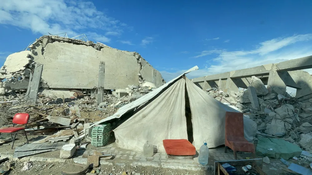 A canvas tent with a red cushion at its door, pitched among the concrete rubble of a bombed building in Gaza