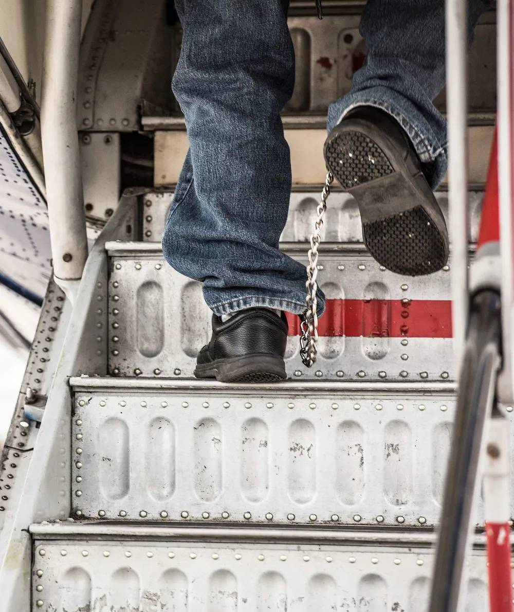 A deportee with a chain between his ankles climbs the steps of an ICE removal flight