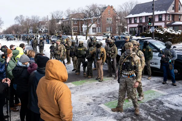 A line of masked, armed federal agents in camouflage facing a crowd of residents filming them on a snowy Minneapolis street