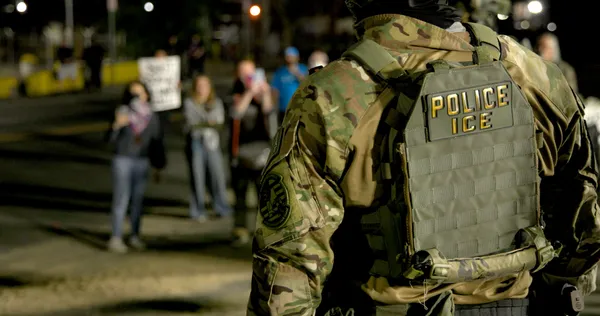 An armed ICE officer in a vest marked Police ICE faces protesters at night outside the Delaney Hall detention centre in Newark