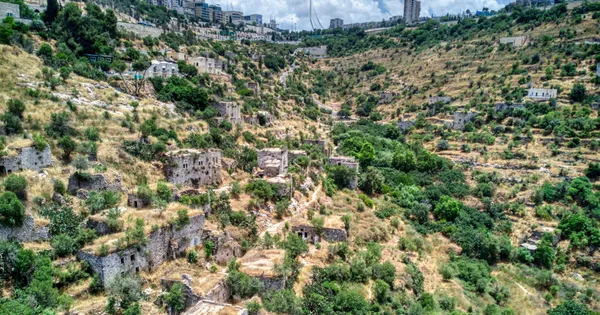 The stone houses of Lifta on terraced slopes, with Jerusalem's high-rise buildings above the valley