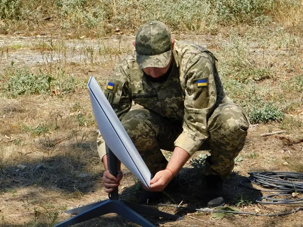 A Ukrainian soldier in camouflage kneels to set up a Starlink satellite dish in a dry field