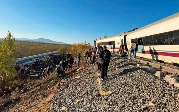 A derailed high-speed train on its side on an embankment with people standing on the track beside it